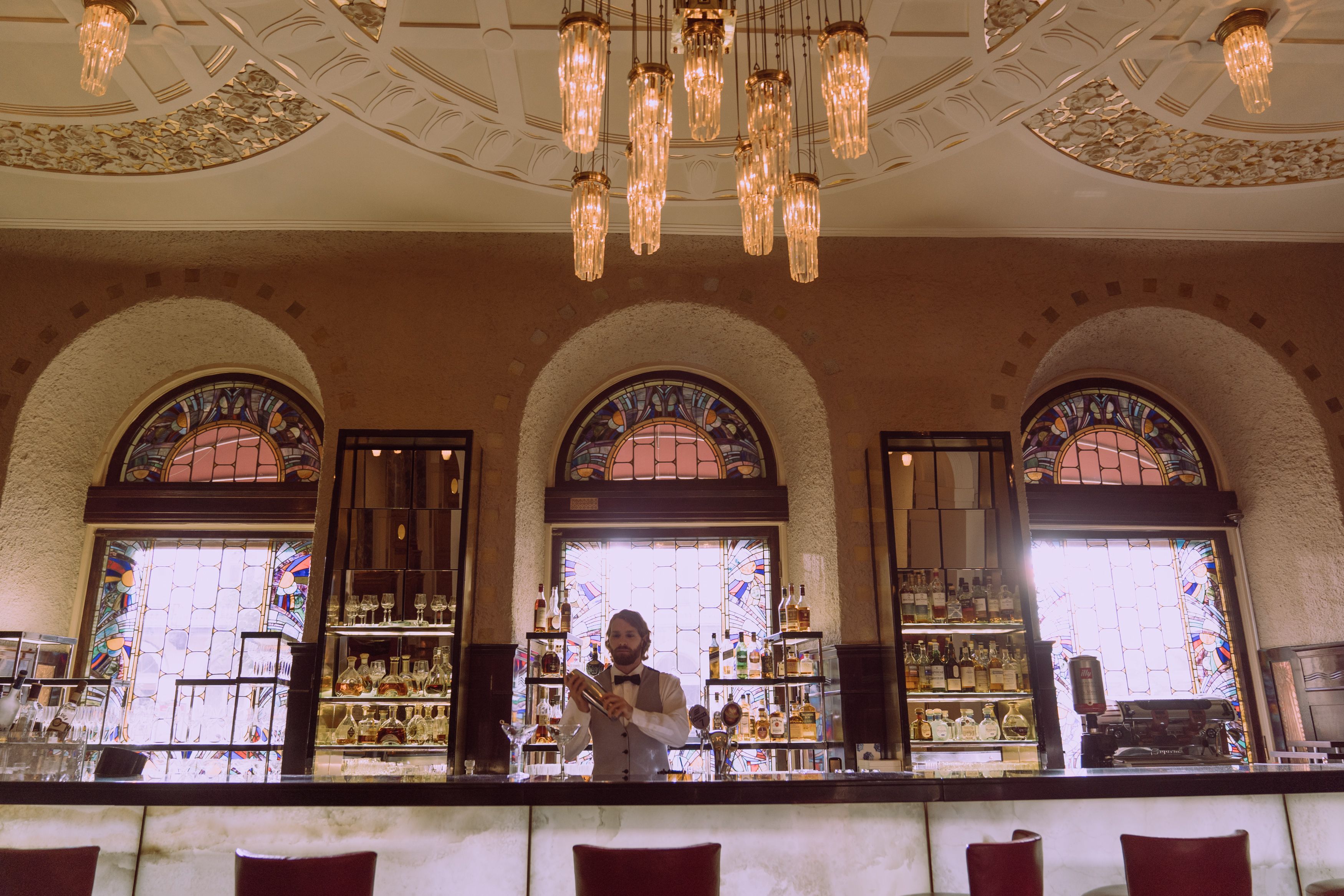 A sophisticated bar interior with chandelier lighting and a bartender serving drinks.
