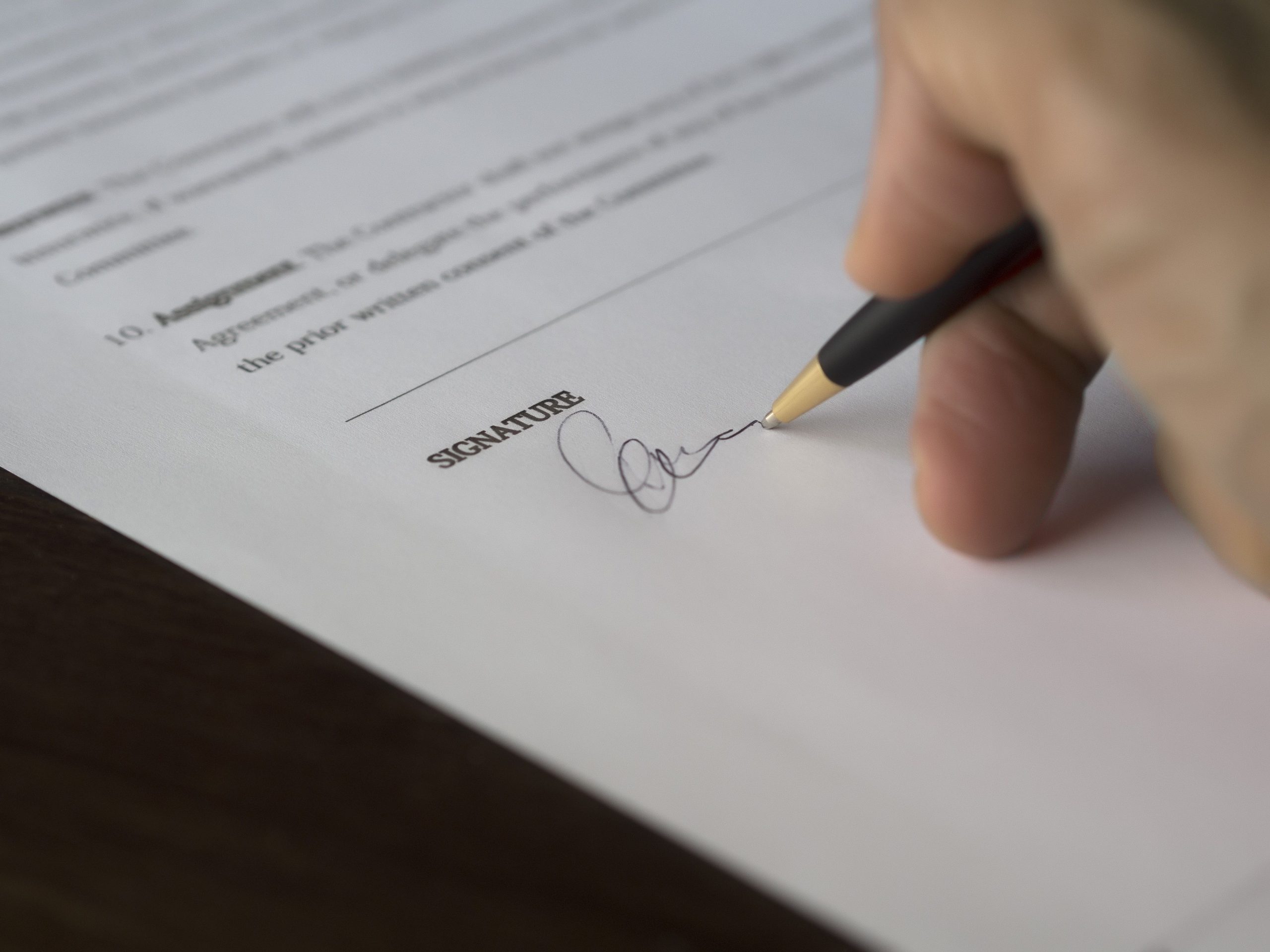 A hand signs a formal contract with a pen on a wooden desk